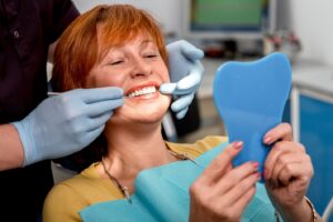 Older patient with dental implants smiling into handheld mirror in dentist’s chair.
